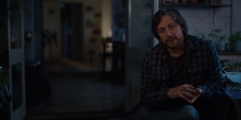 Movie still from “Together” (2021), directed by Justin Martin – A man sitting on a table in a dark room; Medium shot, Over the shoulder angle