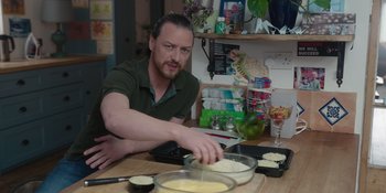 Movie still from “Together” (2021), directed by Justin Martin – A man in a green t - shirt is preparing food; Medium shot, Over the shoulder angle