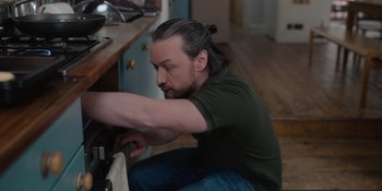 Movie still from “Together” (2021), directed by Justin Martin – A man with long black hair is fixing a stove; Medium shot, High angle