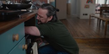 Movie still from “Together” (2021), directed by Justin Martin – A man with long black hair is leaning against a counter; Medium shot, High angle
