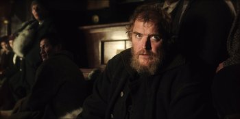 Movie still from “Togo” (2019), directed by Ericson Core – A man with a long beard sitting on a couch; Close Up shot, Over the shoulder angle