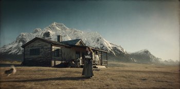 Movie still from “Togo” (2019), directed by Ericson Core – A woman standing in front of a house in a field; Wide shot, Low angle