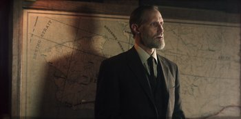 Movie still from “Togo” (2019), directed by Ericson Core – A man wearing a suit and tie standing in front of a map; Medium shot, Low angle