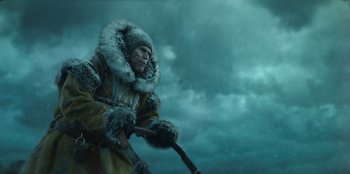 Movie still from “Togo” (2019), directed by Ericson Core – A man in a fur coat holding a stick in the snow; Medium shot, Low angle