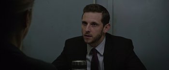 Movie still from “Without Remorse” (2021), directed by Stefano Sollima – A man wearing a suit and tie sitting at a table with a drink; Close Up shot, Over the shoulder angle