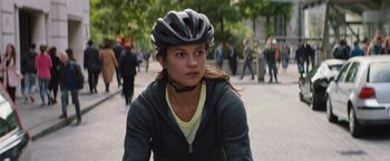 Movie still from “Tomb Raider” (2018), directed by Roar Uthaug – A woman wearing a bicycle helmet while riding down a street; Close Up shot, Over the shoulder angle