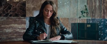 Movie still from “Tomb Raider” (2018), directed by Roar Uthaug – A woman sitting at a table writing on a piece of paper; Medium shot, High angle