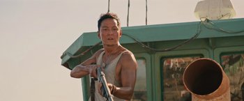 Movie still from “Tomb Raider” (2018), directed by Roar Uthaug – A man holding a gun while standing in front of a boat; Medium shot, Low angle