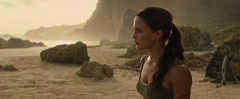 Movie still from “Tomb Raider” (2018), directed by Roar Uthaug – A woman standing on top of a sandy beach near rocks; Close Up shot, Over the shoulder angle