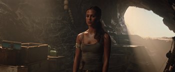 Movie still from “Tomb Raider” (2018), directed by Roar Uthaug – A woman standing in a dark room wearing a gray tank top; Medium shot, Over the shoulder angle