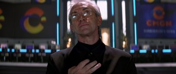 Movie still from “Tomorrow Never Dies” (1997), directed by Roger Spottiswoode – An older man in a black shirt is holding his hand over his heart; Close Up shot, Low angle