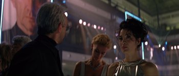 Movie still from “Tomorrow Never Dies” (1997), directed by Roger Spottiswoode – A man and two women standing next to each other in front of lights; Close Up shot, Over the shoulder angle