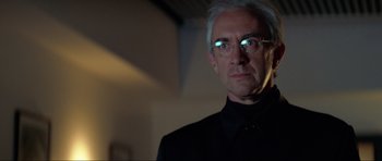 Movie still from “Tomorrow Never Dies” (1997), directed by Roger Spottiswoode – An older man wearing glasses and a black shirt; Close Up shot, Low angle