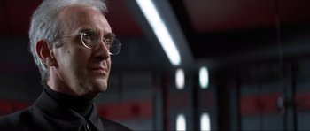 Movie still from “Tomorrow Never Dies” (1997), directed by Roger Spottiswoode – An older man wearing glasses and a black suit; Close Up shot, Low angle