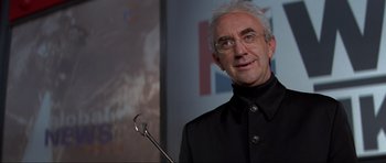 Movie still from “Tomorrow Never Dies” (1997), directed by Roger Spottiswoode – An older man in a black jacket is holding a microphone; Close Up shot, Low angle