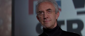 Movie still from “Tomorrow Never Dies” (1997), directed by Roger Spottiswoode – An older man with white hair wearing glasses and a black shirt; Close Up shot, Low angle