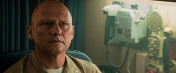 Movie still from “Top Gun” (1986), directed by Tony Scott – A bald man in a tan shirt is in front of a phone; Close Up shot, Low angle