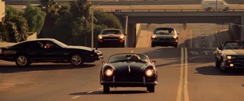 Movie still from “Top Gun” (1986), directed by Tony Scott – An old car driving down a street with cars on the side of the road; Wide shot, High angle