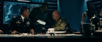 Movie still from “Transformers: Dark of the Moon” (2011), directed by Michael Bay – A man in a military uniform is holding a book while another man looks on; Medium shot, Over the shoulder angle