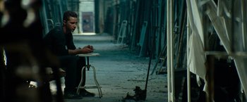 Movie still from “Transformers: Revenge of the Fallen” (2009), directed by Michael Bay – A man sitting on top of a stool next to a table; Wide shot, High angle