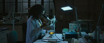 Movie still from “Transformers: Rise of the Beasts” (2023), directed by Steven Caple Jr. – A woman looking through a microscope in a room; Medium shot, High angle