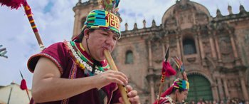 Movie still from “Transformers: Rise of the Beasts” (2023), directed by Steven Caple Jr. – A man wearing a colorful hat playing an instrument in front of a building; Medium shot, Low angle