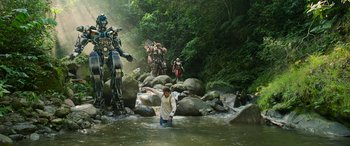 Movie still from “Transformers: Rise of the Beasts” (2023), directed by Steven Caple Jr. – A man standing in a river surrounded by giant robots; Extreme Wide shot, Low angle