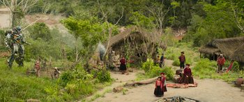 Movie still from “Transformers: Rise of the Beasts” (2023), directed by Steven Caple Jr. – A group of people standing in the dirt near a hut; Extreme Wide shot, Low angle