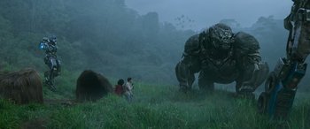Movie still from “Transformers: Rise of the Beasts” (2023), directed by Steven Caple Jr. – Two people are standing in the grass near a giant creature; Extreme Wide shot, Low angle