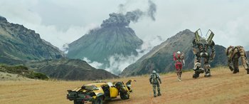 Movie still from “Transformers: Rise of the Beasts” (2023), directed by Steven Caple Jr. – An image of a man standing next to a car in a field; Extreme Wide shot, Low angle