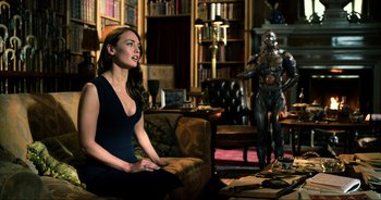 Movie still from “Transformers: The Last Knight” (2017), directed by Michael Bay – A woman sitting on a couch in a living room next to a robot; Medium shot, Over the shoulder angle