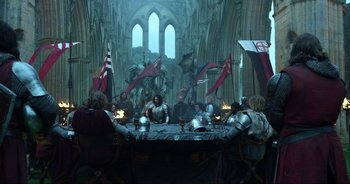 Movie still from “Transformers: The Last Knight” (2017), directed by Michael Bay – A group of people sitting around a table with flags; Wide shot, Over the shoulder angle