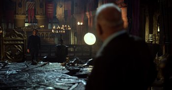 Movie still from “Transformers: The Last Knight” (2017), directed by Michael Bay – An older man sitting at a table in a dark room; Wide shot, Over the shoulder angle