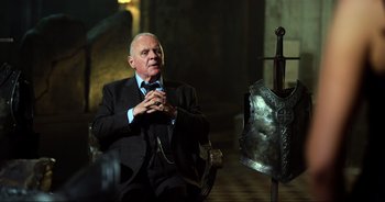 Movie still from “Transformers: The Last Knight” (2017), directed by Michael Bay – An older man in a suit and tie sitting next to a metal shield; Medium shot, Over the shoulder angle