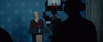 Movie still from “Troll” (2022), directed by Roar Uthaug – A woman standing at a podium in front of a video camera; Medium shot, High angle