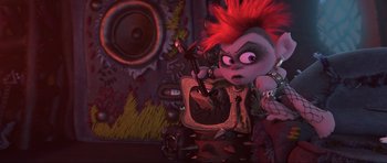 Movie still from “Trolls World Tour” (2020), directed by Walt Dohrn – A cartoon character with a red mohawk hair style is sitting on a television set; Medium shot, High angle