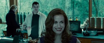 Movie still from “Twilight” (2008), directed by Catherine Hardwicke – A woman with red curly hair smiles at the camera while a man stands in the background; Medium shot, Over the shoulder angle