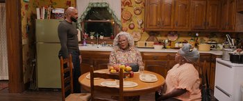 Movie still from “Tyler Perry's A Madea Homecoming” (2022), directed by Tyler Perry – A woman sitting at a table in a kitchen with other people; Wide shot, High angle