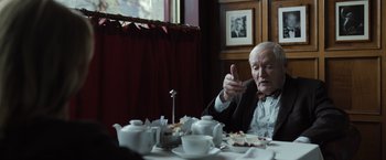 Movie still from “Tár” (2022), directed by Todd Field – An older man sitting at a table with a cup of tea; Medium shot, Over the shoulder angle