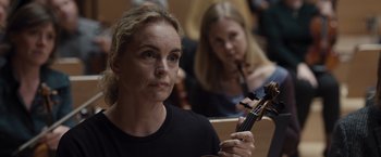 Movie still from “Tár” (2022), directed by Todd Field – A woman is holding a violin in front of other people; Close Up shot, Over the shoulder angle