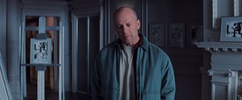 Movie still from “Unbreakable” (2000), directed by M. Night Shyamalan – A bald man in a blue jacket is standing in a dark room; Medium shot, Over the shoulder angle