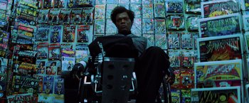 Movie still from “Unbreakable” (2000), directed by M. Night Shyamalan – A man sitting in a wheelchair in front of a wall of comic books; Medium shot, Low angle