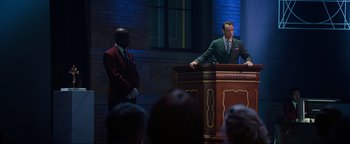 Movie still from “Uncharted” (2022), directed by Ruben Fleischer – A man standing at a podium in front of a crowd; Wide shot, Over the shoulder angle