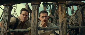 Movie still from “Uncharted” (2022), directed by Ruben Fleischer – A man standing next to another man in a wooden structure; Medium shot, Low angle