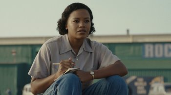 Movie still from “A League of Their Own” (2022), created by Abbi Jacobson – A woman sitting on the ground writing on a piece of paper; Medium shot, Low angle