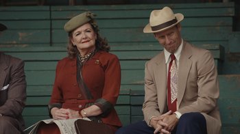 Movie still from “A League of Their Own” (2022), created by Abbi Jacobson – An older woman sitting next to an older man wearing a hat; Medium shot, Over the shoulder angle