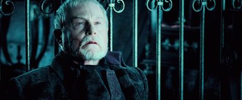 Movie still from “Underworld: Evolution” (2006), directed by Len Wiseman – An older man with a beard and mustache sitting in front of a metal fence; Close Up shot, Over the shoulder angle