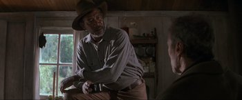 Movie still from “Unforgiven” (1992), directed by Clint Eastwood – An older man wearing a cowboy hat while holding a gun; Medium shot, Over the shoulder angle