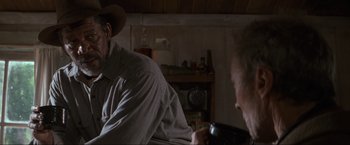 Movie still from “Unforgiven” (1992), directed by Clint Eastwood – A man sitting on top of a couch next to another man holding a tv remote; Medium shot, Over the shoulder angle