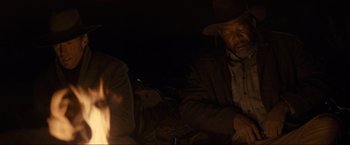 Movie still from “Unforgiven” (1992), directed by Clint Eastwood – A man in a cowboy hat sitting in front of a fire; Medium shot, Over the shoulder angle